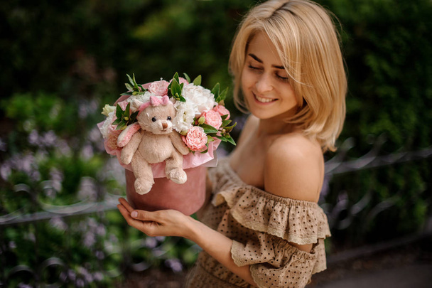 Blonde smiling woman holding a box filled with pink and white flowers decorated with a cute toy - Photo, image