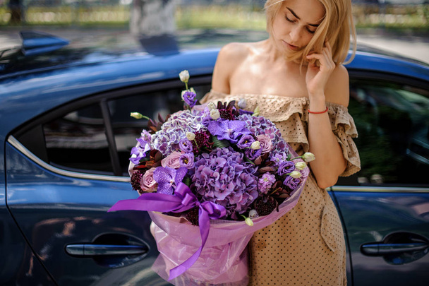 Blonde woman in beige dress holding a big bouquet of flowers in bright and soft purple tones against the blue car - Photo, Image