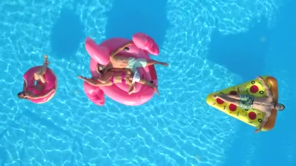 Luchtfoto 's Top Down: Lachende meisjes en jongens in zwemkleding op leuke opblaasbare pizza, donut en flamingo drijvend op water. Gelukkige vrienden genieten op roze flamingo, donut en pizza drijft in het zwembad - Video