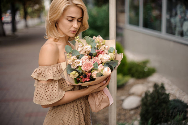 Romantic blonde woman in dress holding a nude beige bouquet of flowers wrapped in craft paper - Fotó, kép