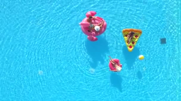 Luchtfoto Top Down Fit meisjes en jongens in zwempakken liggen op leuke opblaasbare pizza, donut en flamingo drijvend op water. Vrienden op vakantie genietend van roze flamingo, donut en pizza drijvers in het zwembad - Video