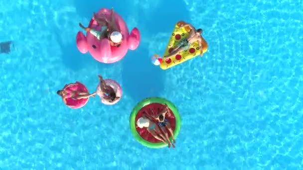 Luchtfoto Top Down lachende meisjes en jongens in zwemkleding opleggen leuke opblaasbare pizza, donut en flamingo drijvend op het water. Happy vrienden genieten op roze flamingo, drijft donut en de pizza in zwembad - Video