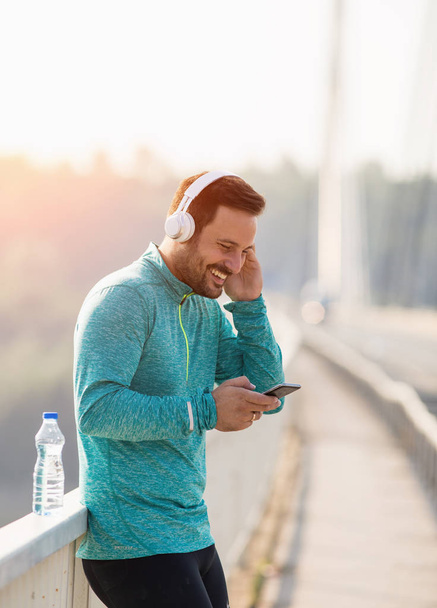 Happy handsome man taking break after running and listening music on bridge - Фото, изображение