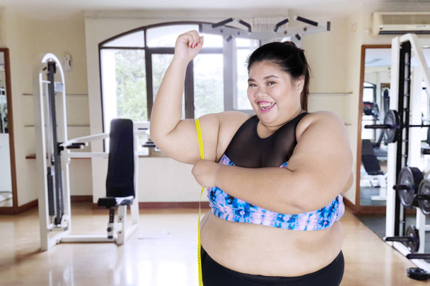 Picture of overweight woman looks happy while measuring her bicep and standing in the gym center - Foto, Imagen