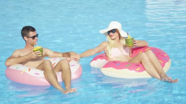 Αργή κίνηση: Νεαρό ζευγάρι πίνοντας κοκτέιλ ανανά και χαλαρωτικό διασκέδαση ροζ ντόνατ floaties στην πισίνα. Νεαρός άνδρας και γυναίκα αγάπη χαλαρώνει στο φουσκωτό ντόνατ επιπλέει στις ηλιόλουστες καλοκαιρινές διακοπές - Πλάνα, βίντεο