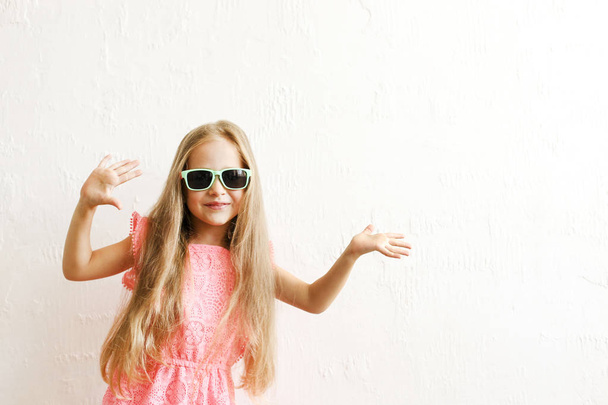 Little girl w/ long golden hair in fashionable casual outfit, wearing sunglasses & smiling, laughing, making funny faces. Five years old daughter in pink dress posing. Background, close up, copy space - Foto, Imagem