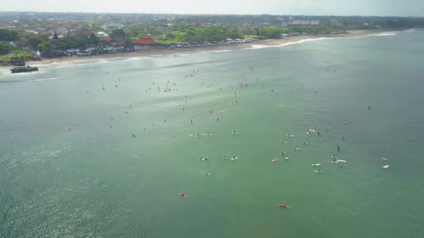 AERIAL: Flying above crowded surfspot in Canggu village. Oceanfront villas, hotels, bungalows and houses in tourist resort on sandy beach in Bali. People in lineup waiting breaking waves in calm ocean - Footage, Video