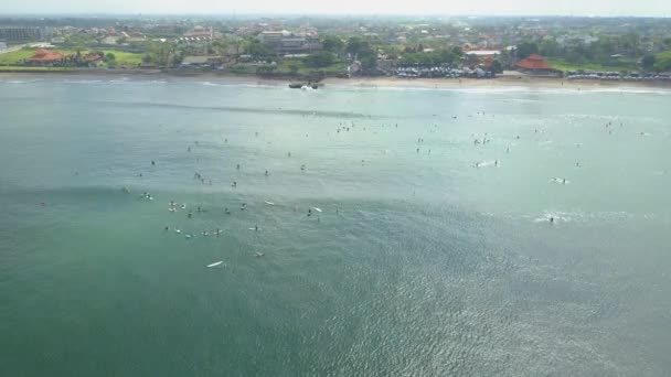 AERIAL: Flying above crowded surfspot in Canggu village. Oceanfront villas, hotels, bungalows and houses in tourist resort on sandy beach in Bali. People in lineup relaxing on surfboards in calm ocean - Footage, Video
