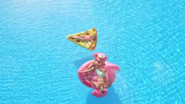 Aerial Top Down Lens Flare: Młodzi mężczyźni i kobiety w strojach kąpielowych kładący się na zabawnej nadmuchiwanej pizzy i flamingu unoszącym się na wodzie. Szczęśliwi przyjaciele na wakacjach korzystających z różowego flaminga i pizzy pływa w basenie - Materiał filmowy, wideo
