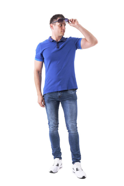 Homem adulto macho jovem confiante em camisa de pólo azul segurando e olhando para longe sob óculos de sol. Corpo inteiro isolado em fundo branco
. - Foto, Imagem