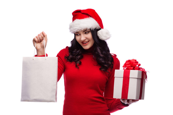 Beautiful brunette woman wearing santa hat and holding gift box and shopping bag. Isolated on white background - Фото, изображение