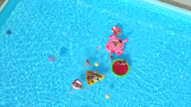 AERIAL: Cheerful friends having water fight on colorful floaties at pool party. Playful guys and girls splashing water and taking selfies on inflatable pizza, doughnut, flamingo and watermelon floats - Felvétel, videó