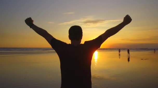 SLOW MOTION, CLOSE UP, SILHOUETTE: Excited traveler man standing on stunning sandy beach watching splashing ocean waves and raising hands at golden sunrise. Happy tourist guy extending arms at sunset - Footage, Video