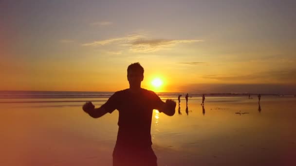 Lassú mozgás, szorosan fel, lencse fényfolt: Izgatott fiatal férfi ujjongott a felemelt álmodozó homokos strandon a mágikus narancssárga naplemente. Boldog emelés a srác kezét a levegőben az izgalom. Fő ünnepek - Felvétel, videó