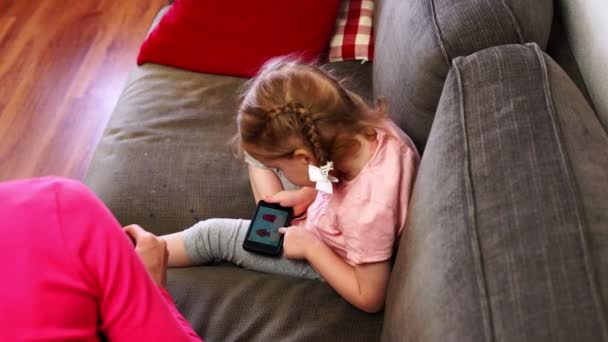 Little girl plays on her smartphone while her mother feeds her a spoon - Filmagem, Vídeo