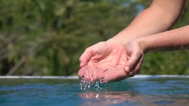 Zwolnionym tempie Close Up kaukaski kobiece ręce grać z wody, łowienie to palcami & leje do nieskończoności puli na rajskiej wyspie. Człowieka z palmami nabiera wody, krople na powierzchni wody - Materiał filmowy, wideo