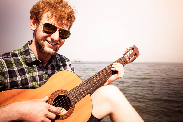 Happy young man hipster playing guitar by sea ocean water. Handsome guy in sunglasses relaxing on summer vacation. - Foto, imagen