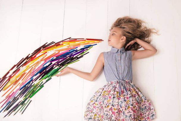 Een leuk jong meisje in een jurk met vlinders ligt op de grond en blaast een stroom van gekleurde potloden - Foto, afbeelding