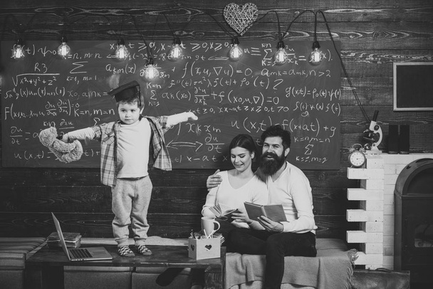 Support concept. Kid holds teddy bear and performing. Boy presenting his knowledge to mom and dad. Parents listening their son, chalkboard on background. Smart child in graduate cap like to perform - Фото, зображення