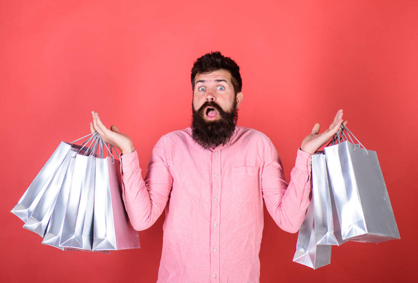 Guy shopping on sales season with discounts. Man with beard and mustache holds shopping bags, red background. Hipster on shocked face is shopping addicted or shopaholic. Sale and discount concept - 写真・画像