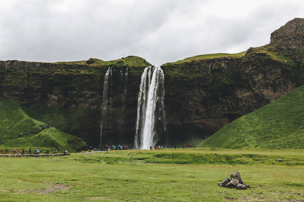 Island - 20 června 2018: krásný vodopád Seljalandsfoss a turisté za zatažené obloze  - Fotografie, Obrázek