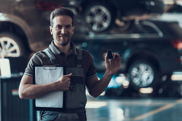 Auto Mechanic Holding Clipboard and Car Keys. Repair service. Handsome Serious Caucasian Man in Grey Uniform Standing in Front of Black Car and Finished Checkup of Service Order Working in Garage - Photo, Image