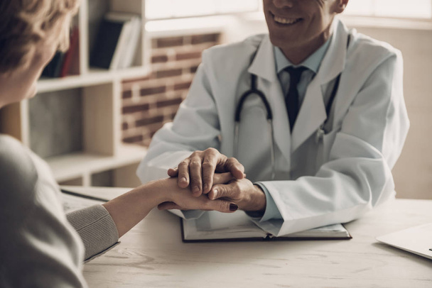 Close-up of Smiling Doctor Holding Patients Hand. Confident Medical Worker Sitting at Table in Cabinet and Making Conversation about Diagnosis with Hopefull Expression. Medical Issue Concept - 写真・画像