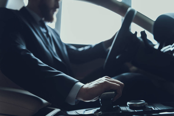 Close-up Of Male Hand Change Gear and Driving Car. Caucasian Person Wearing Black Suit Posing in Auto Using Car Transmission. Driving Automobile with Automatic Gears Concept - Foto, afbeelding