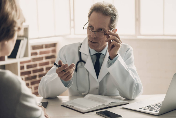 Doctor talking with Patient with Serious Face. Middleaged Confident Medical Worker Sitting at Table in Cabinet and Making Conversation about Diagnosis. Medical issue concept - Photo, Image