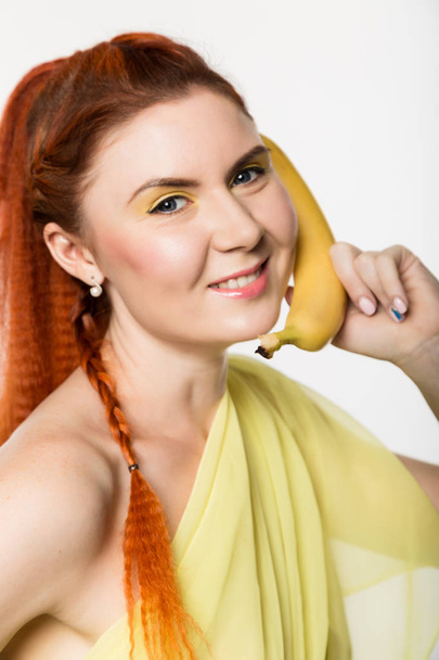 Giovane donna rossa che tiene banana vicino al viso come uno smartphone su uno sfondo bianco
. - Foto, immagini