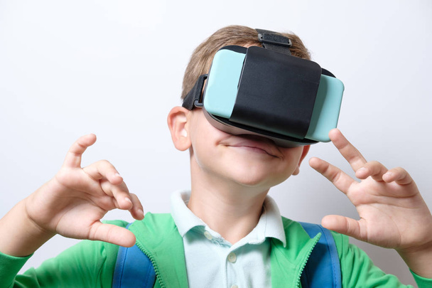 Cute schoolboy with blue backpack wearing virtual reality helmet with arms stretched forward on a white background - Foto, Imagem