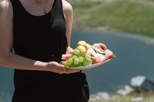 Young woman in a black dress holds a plate of fruit: pears, watermelon, grapes and figs at picnic in the mountains Montenegro. - Photo, image