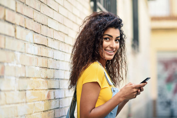 Jonge Arabische vrouw texting met haar Smartphone op de muur van de stad. Lachende Noord Afrincan meisje in casual kleding met zwart krullend kapsel. - Foto, afbeelding