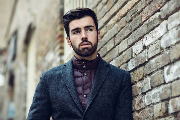 Young bearded man, model of fashion, in urban background wearing british elegant suit. Guy with beard and modern hairstyle in the street. - Photo, Image