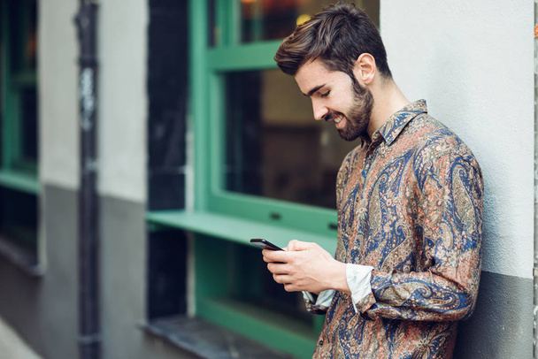 Ein junger lächelnder Mann in lässiger Kleidung blickt auf der Straße auf sein Smartphone. Typ mit Bart und moderner Frisur im urbanen Hintergrund - Foto, Bild