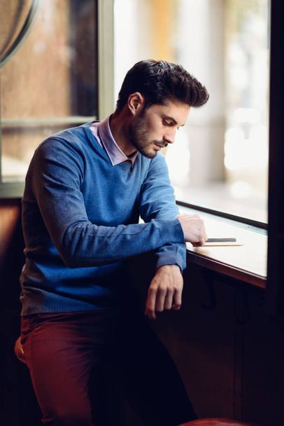 Hombre joven con suéter azul mirando a su teléfono inteligente en un pub moderno. Tipo barbudo con peinado moderno cerca de una ventana
. - Foto, imagen