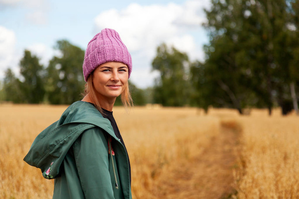 Young dark-haired woman in a knitted pink hat from a natural fur smiling and posing positively on an autumn day against a background of a golden field and a  forest. Village Livestyle - Photo, Image