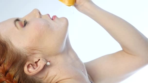 Young woman squeezing orange into the mouth and on your neck on a white background, slow motion. - Metraje, vídeo