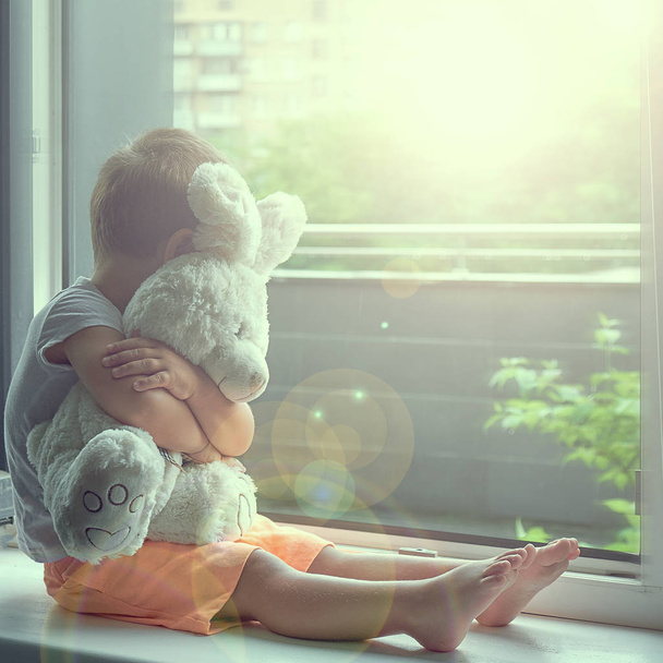 Мальчик двух лет сидит у окна и обнимает игрушечного кролика. дождливая погода, ожидание возвращения папы с работы
 - Фото, изображение