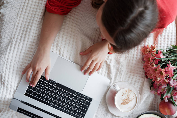 Draufsicht der blonden Frau in rotem Gewand, die am Laptop auf dem Bett arbeitet und vor Fensterhintergrund Kaffee trinkt  - Foto, Bild