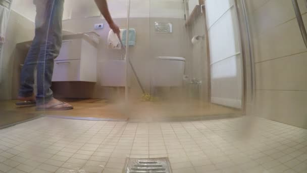 Άνθρωπος με σαγιονάρες και παντελόνι πλένει το πάτωμα στο μπάνιο, άποψη από το ντους που έχει fogged τα ποτήρια με το ζεστό νερό - Πλάνα, βίντεο