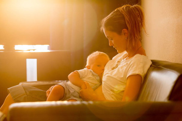 Young mother, breastfeeding on sunset on quiet evening her little baby boy in sunny living room - Foto, afbeelding
