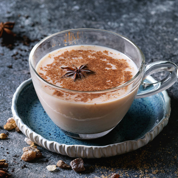 Glass cup of traditional indian masala chai tea on blue saucer with ingredients above. Cinnamon, cardamom, anise, sugar, black tea over dark texture background. Close up. Square image - Fotoğraf, Görsel
