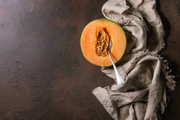 Half of ripe organic Cantaloupe melon with seeds and spoon inside on cloth textile over dark brown texture background. Flat lay, space - Φωτογραφία, εικόνα