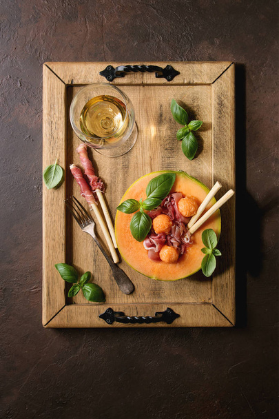 Melon and ham salad served in half of Cantaloupe melon with fresh basil and grissini bread on wooden serving tray over dark brown texture background with glass of white wine. Flat lay, space - Photo, Image