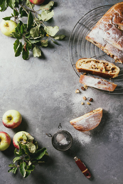Homemade sliced puff pastry apple strudel pie on cooling rack served with ripe fresh apples, branches and sugar powder over grey texture background. Flat lay, space - Foto, Bild