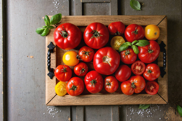 Variety of ripe fresh organic gardening tomatoes different kind and colors with water spot and basil leaves in wood tray over old grey wooden background. Flat lay, space - Photo, Image