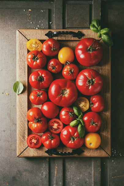 Variety of ripe fresh organic gardening tomatoes different kind and colors with water spot and basil leaves in wood tray over old grey wooden background. Flat lay, space - Photo, image