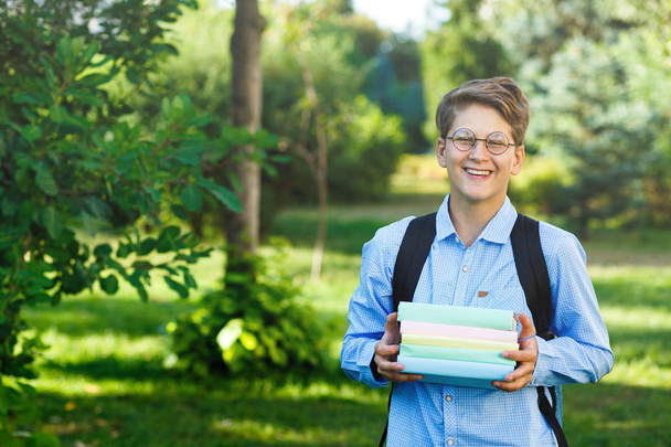 very cute, young boy in round glasses and blue shirt with backpack holds books in the park. Education, back to school concept - Фото, изображение