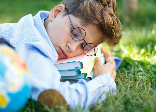 schattige jongen in ronde bril in blauw shirt slaapt op zijn boeken op het gras in het park. Onderwijs, terug naar school concept - Foto, afbeelding